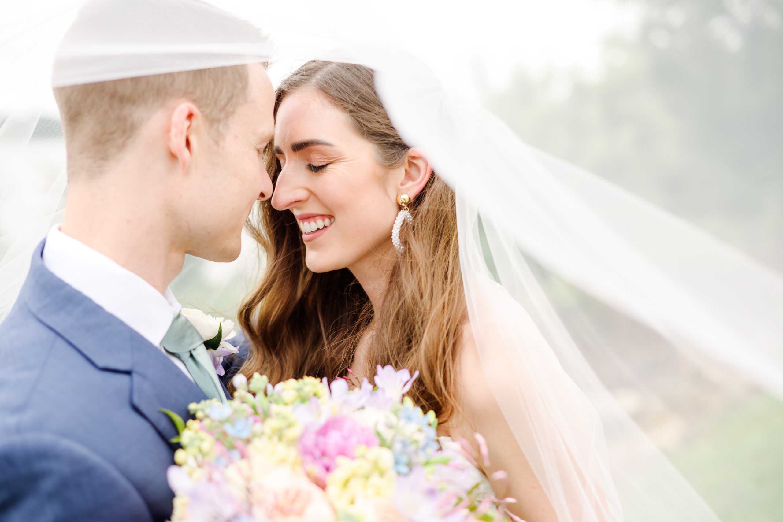 The image shows a close-up of a bride and groom on their wedding day. They are facing each other with their foreheads nearly touching, both smiling happily. The bride is wearing a white veil and has long brown hair. She's also wearing dangling earrings. The groom is wearing a blue suit with a white shirt and a light-colored tie. He has a boutonnière pinned to his lapel. In the foreground, there's a colorful bouquet of flowers, including pink, blue, and yellow blooms. The background is soft and out of focus, giving the image a dreamy, romantic quality. The photo captures an intimate moment between the newlyweds, conveying joy and love on their special day.