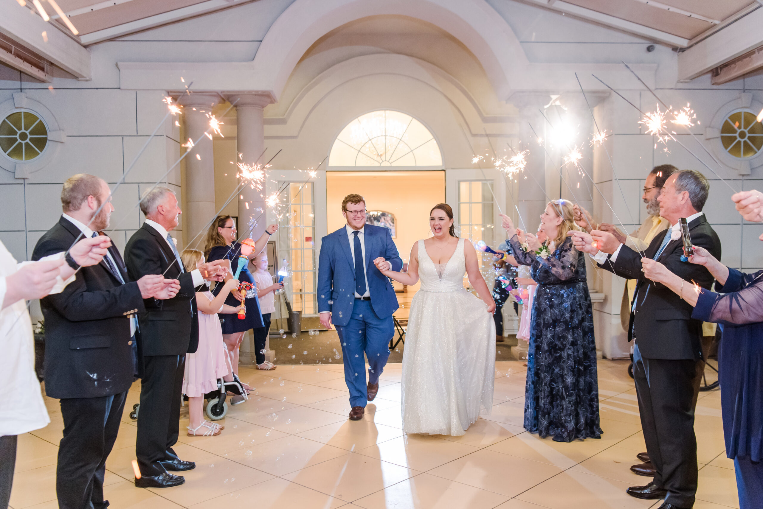 Newlyweds exit ceremony through archway as guests cheer with sparklers, creating a joyful wedding scene in Dallas Ashton Gardens Chapel.