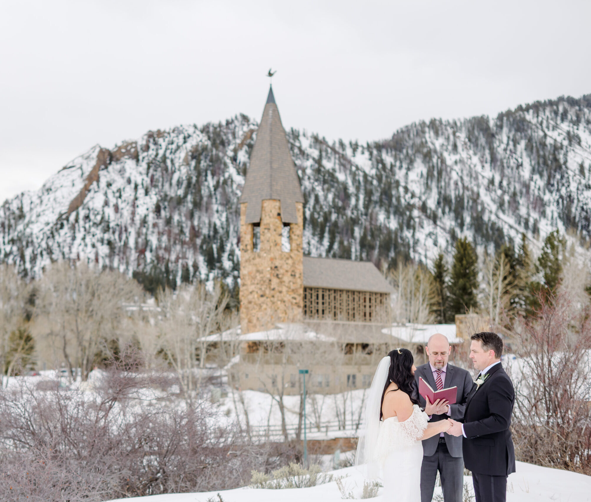 Winter wedding ceremony in front of Aspen Chapel with snow-covered mountains in background.